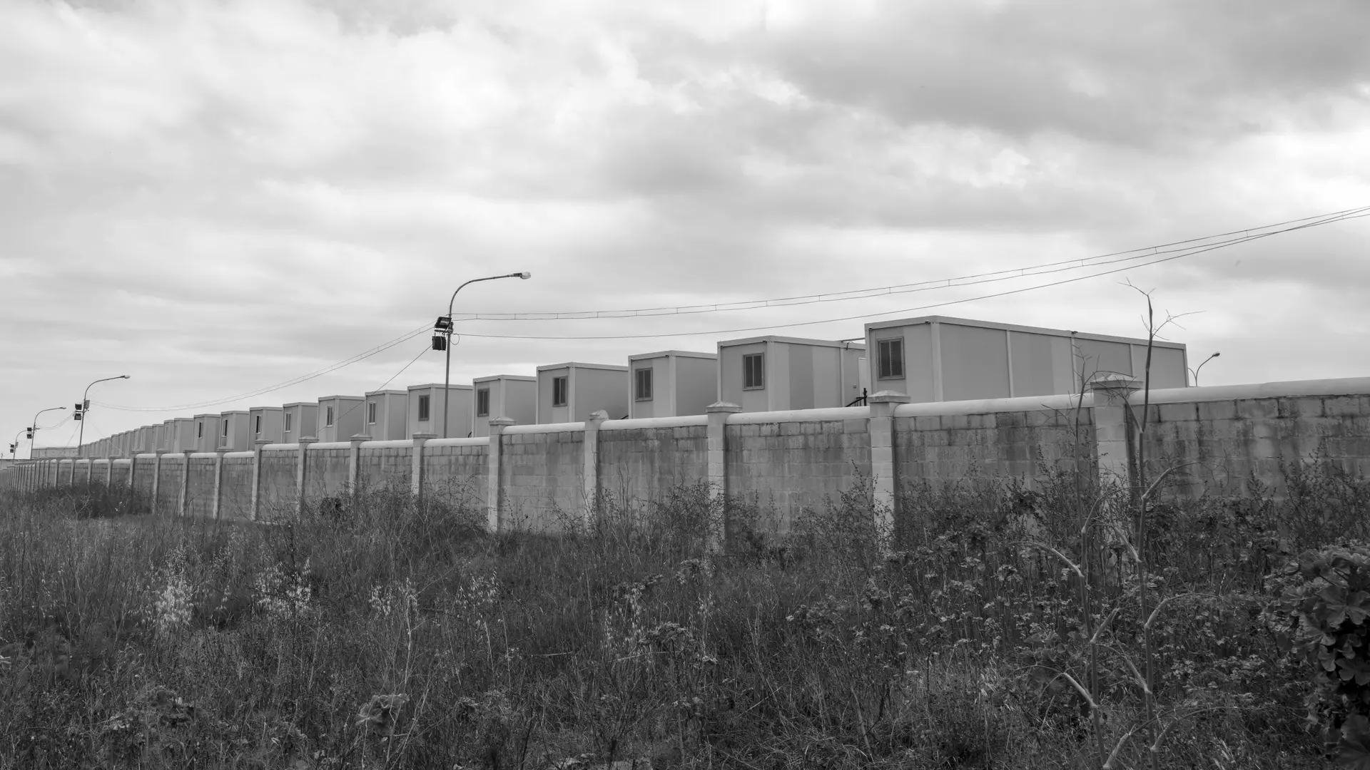 Black and white photo of container houses behind a wall, under a cloudy sky.
