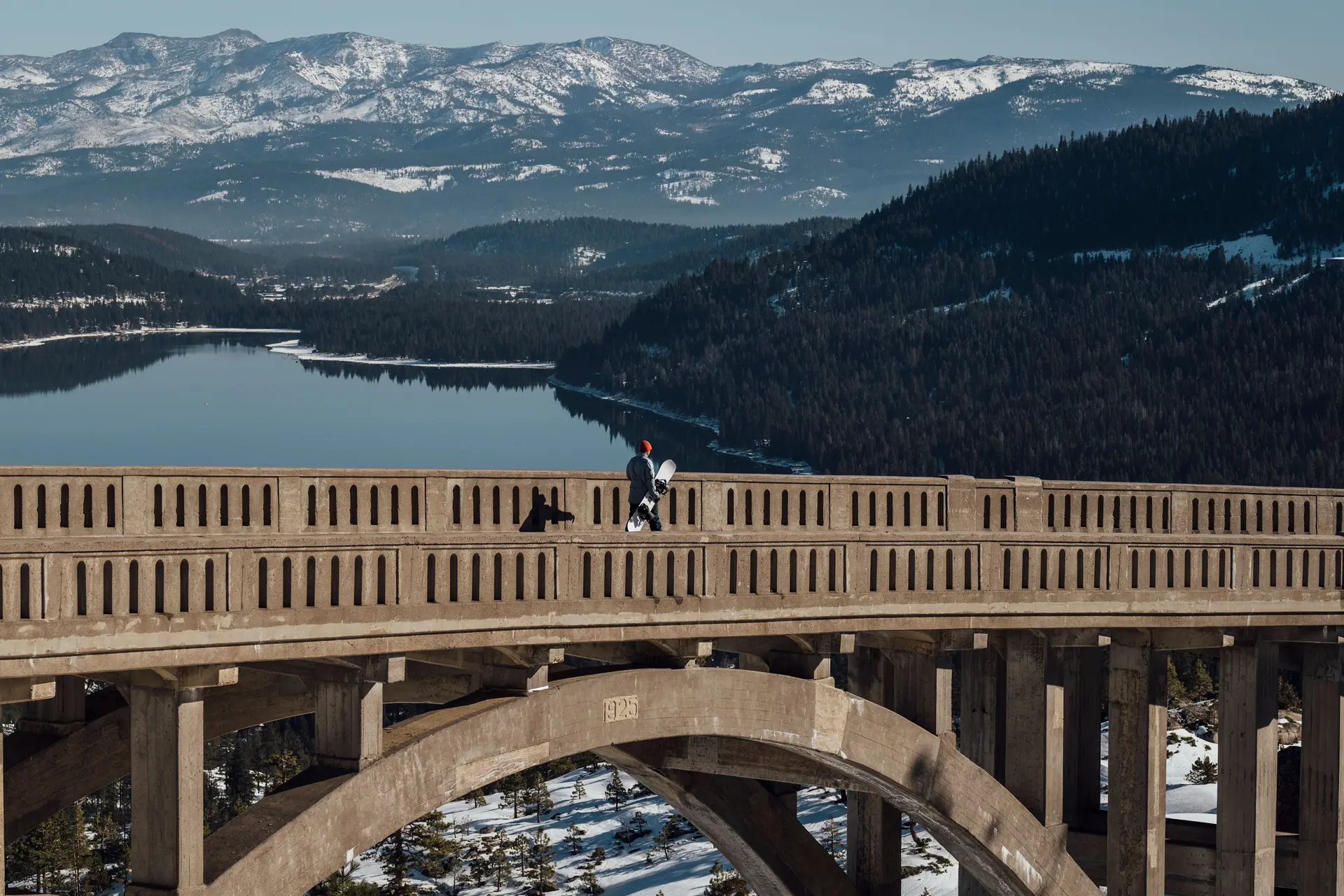 Man with snowboard in hand crosses a large bridge in the mountains with a view of a lake
