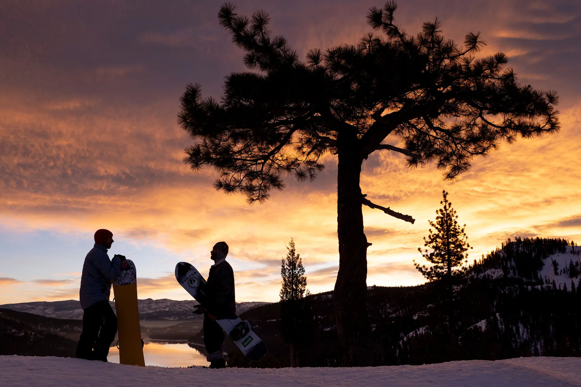 Two snowboarders stand on the mountain in the sunset and talk to each other