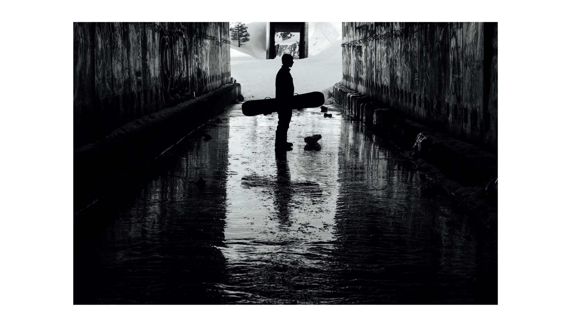 Black and white picture of a snowboarder with snowboard in hand standing in a tunnel