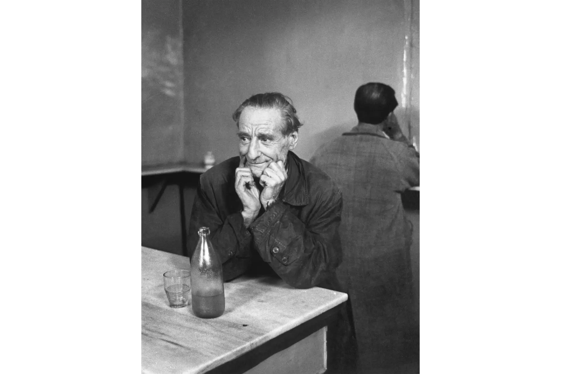 Old man sitting at a table. There is a bottle and a glass on the table