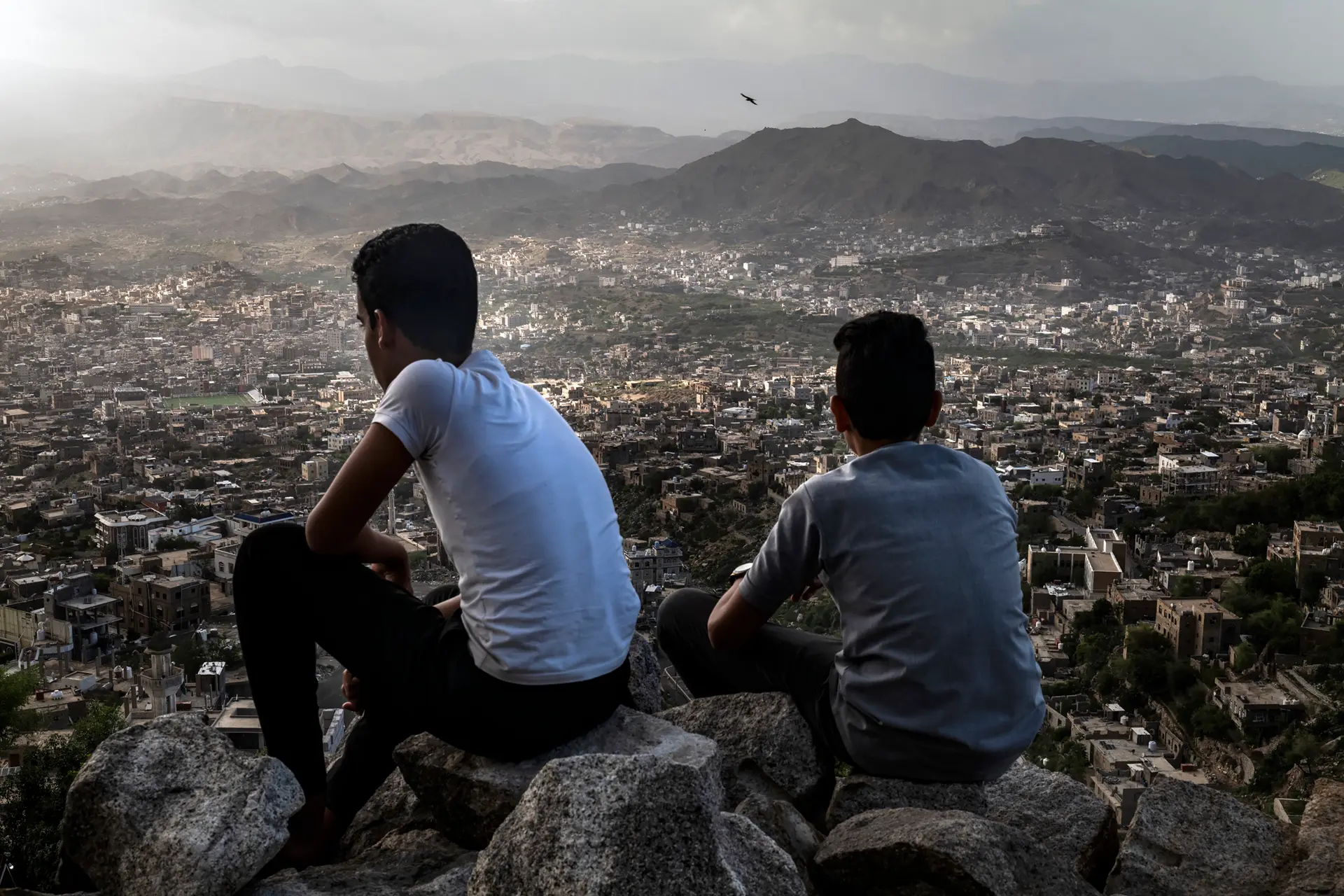 2 boys sit on a mountain and look down on the city of Yemen