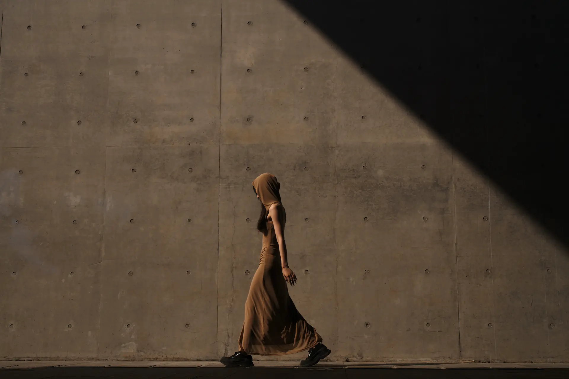 Woman with brown dress, hood and black sneakers walks along a grey concrete wall