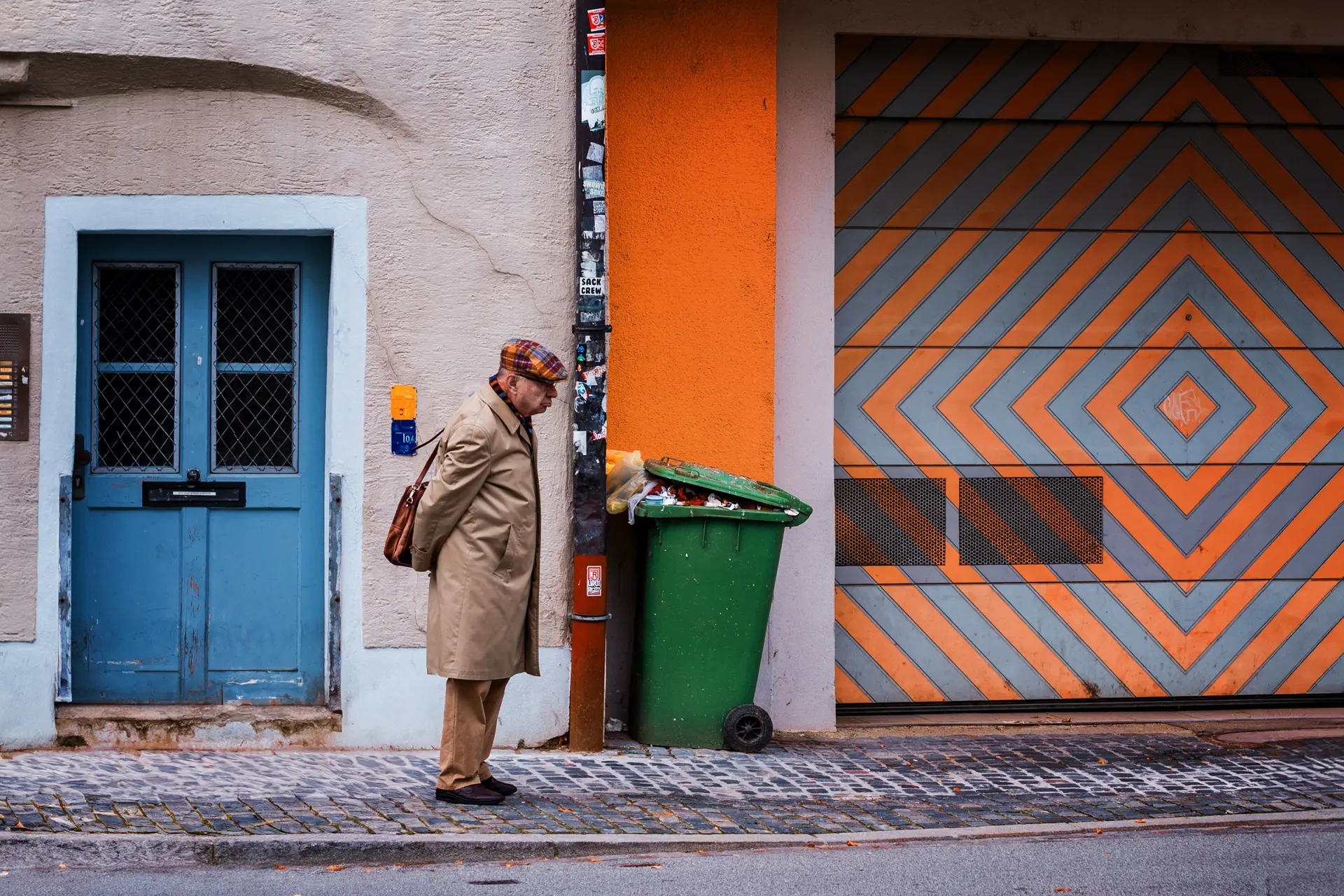 Old man standing between a blue door and a full, overflowing green bin