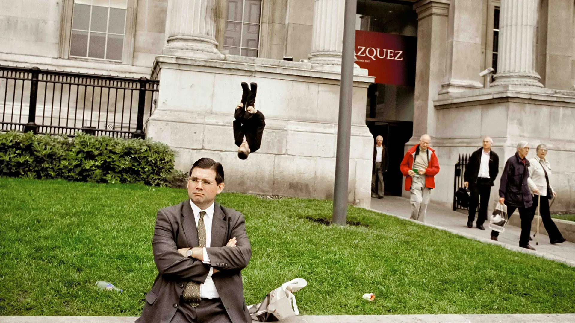 Man in a suit sits on a wall behind him a man in a suit does a somersault