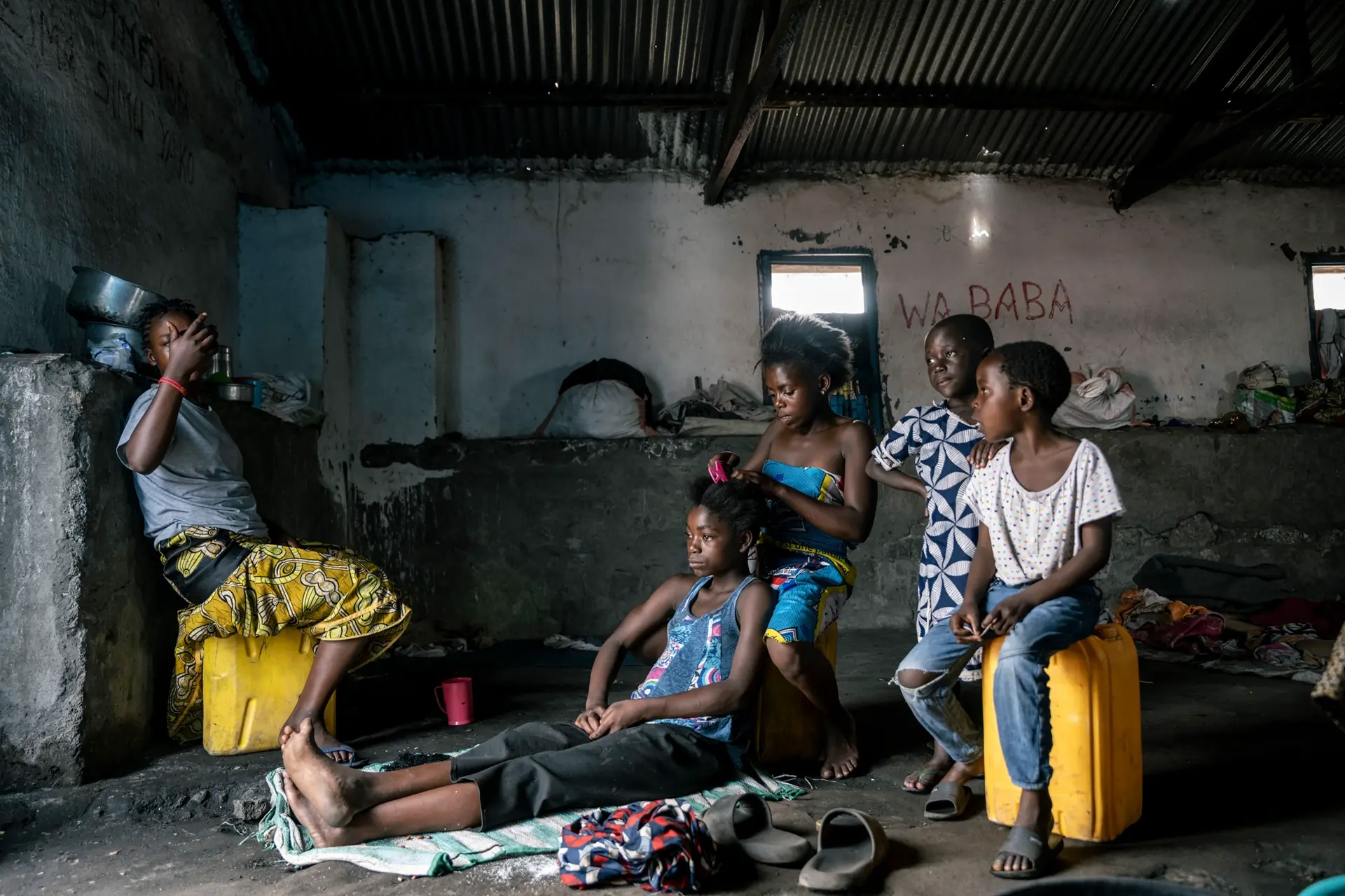 Five children sitting in a run-down hut. One girl braids the other's hair.