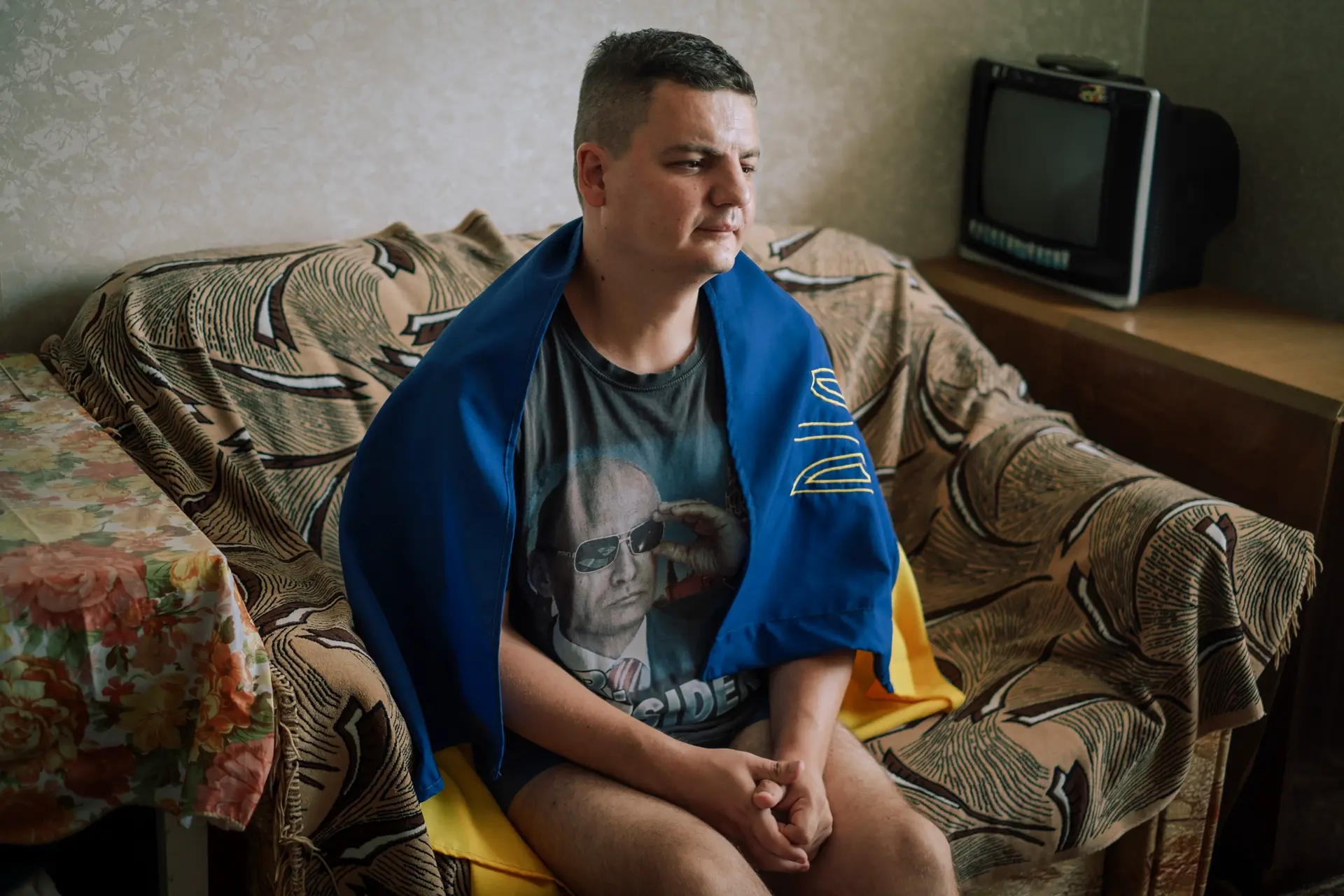 A man sitting sad on his couch with a flag over his shoulders.