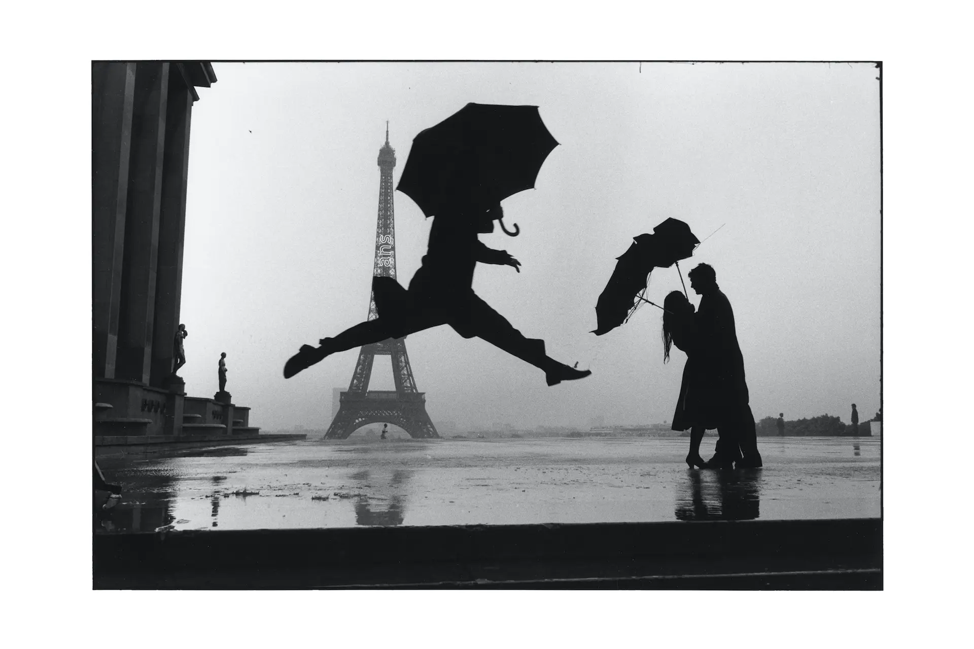 Man with umbrella takes a long jump in the background, the Eiffel Tower and a couple in love can be seen black-and-white photograph Paris, France 1989