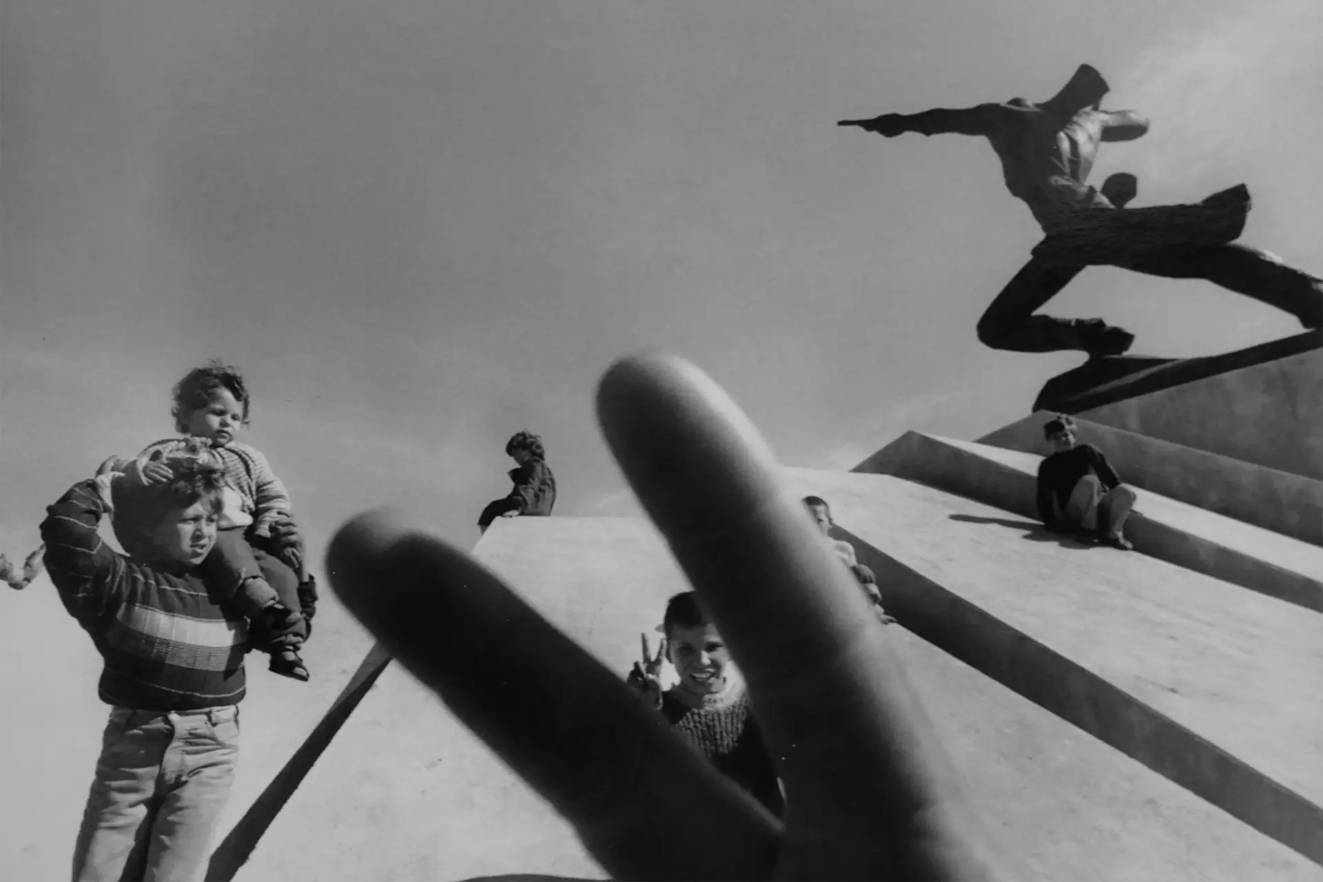 In the foreground fingers in a peace sign in the background, children.