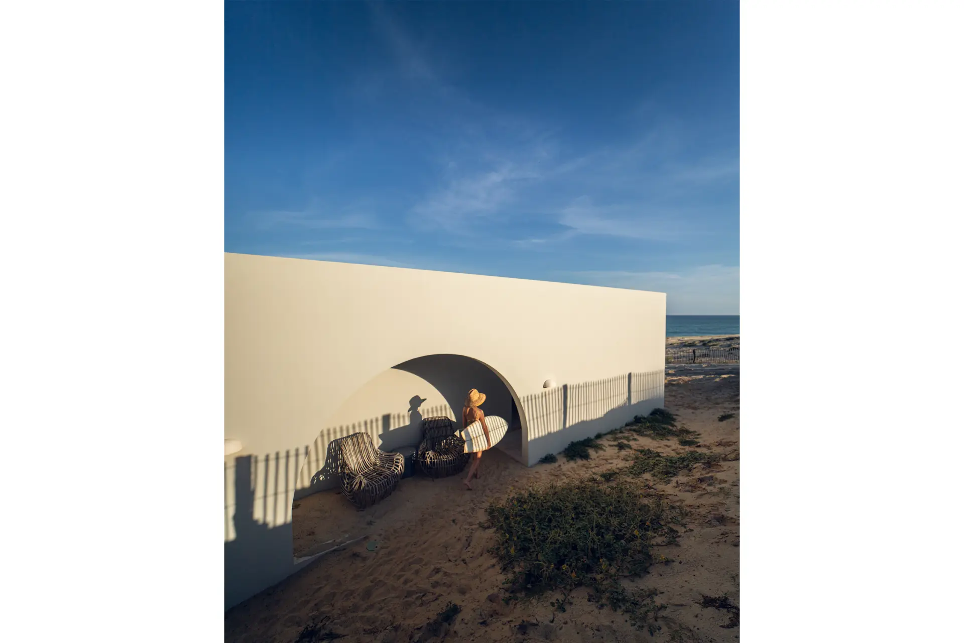 Woman with surfboard under her arm in front of a white building with the sea in the background