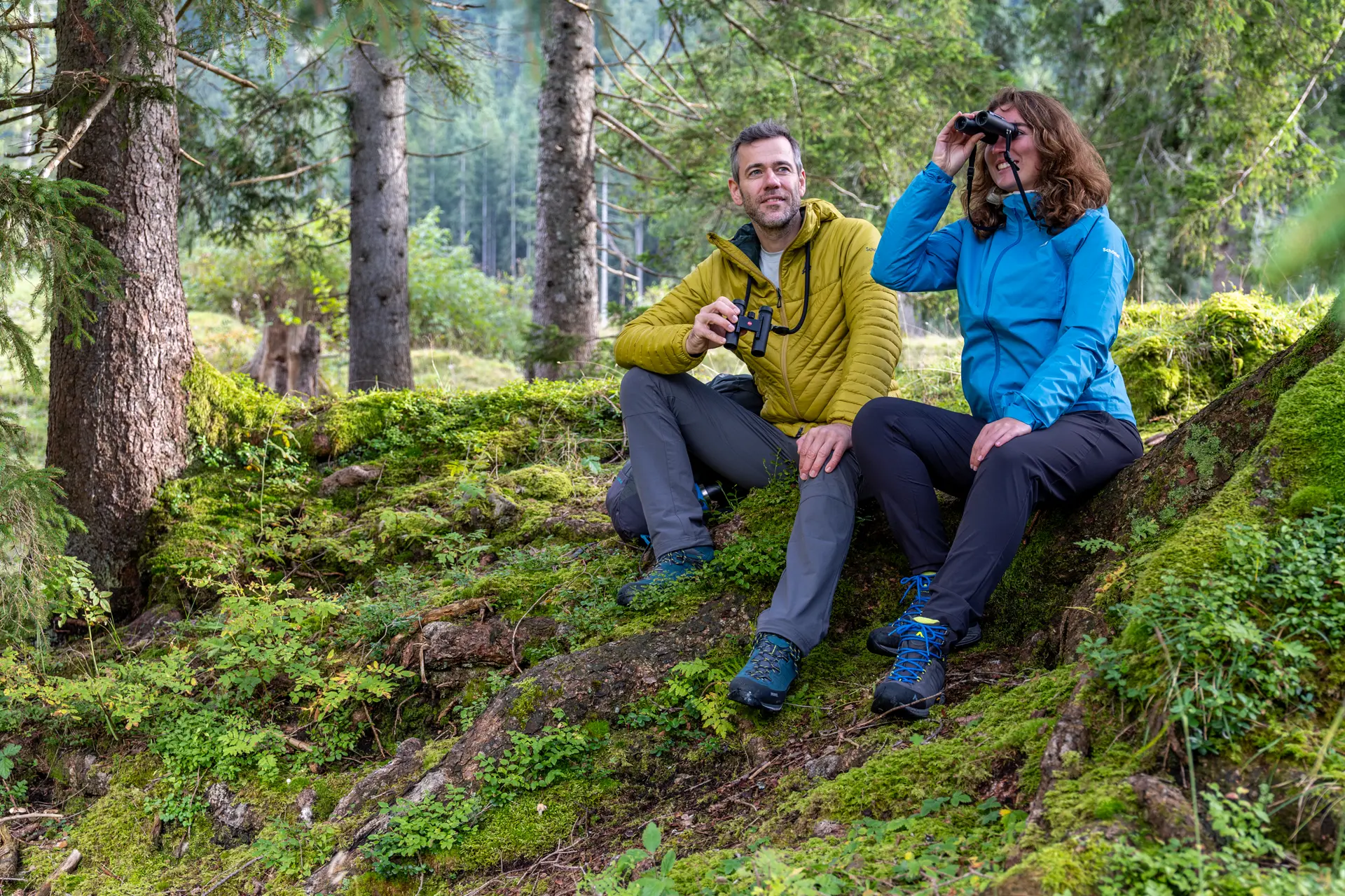 Two hikers are sitting on the ground and looking through their Leica Noctivid Compact