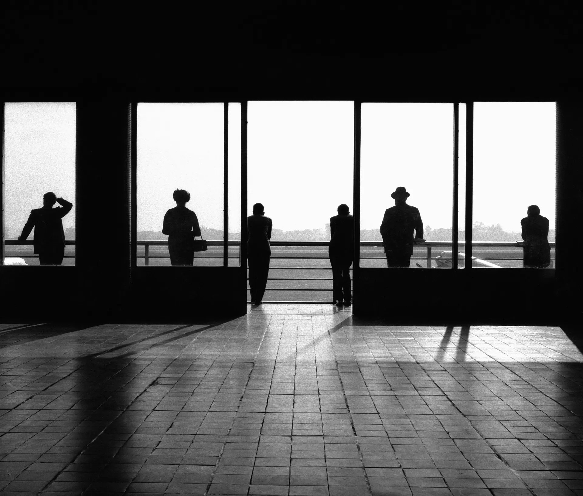 Black and white photograph of people standing in an airport, seen from behind