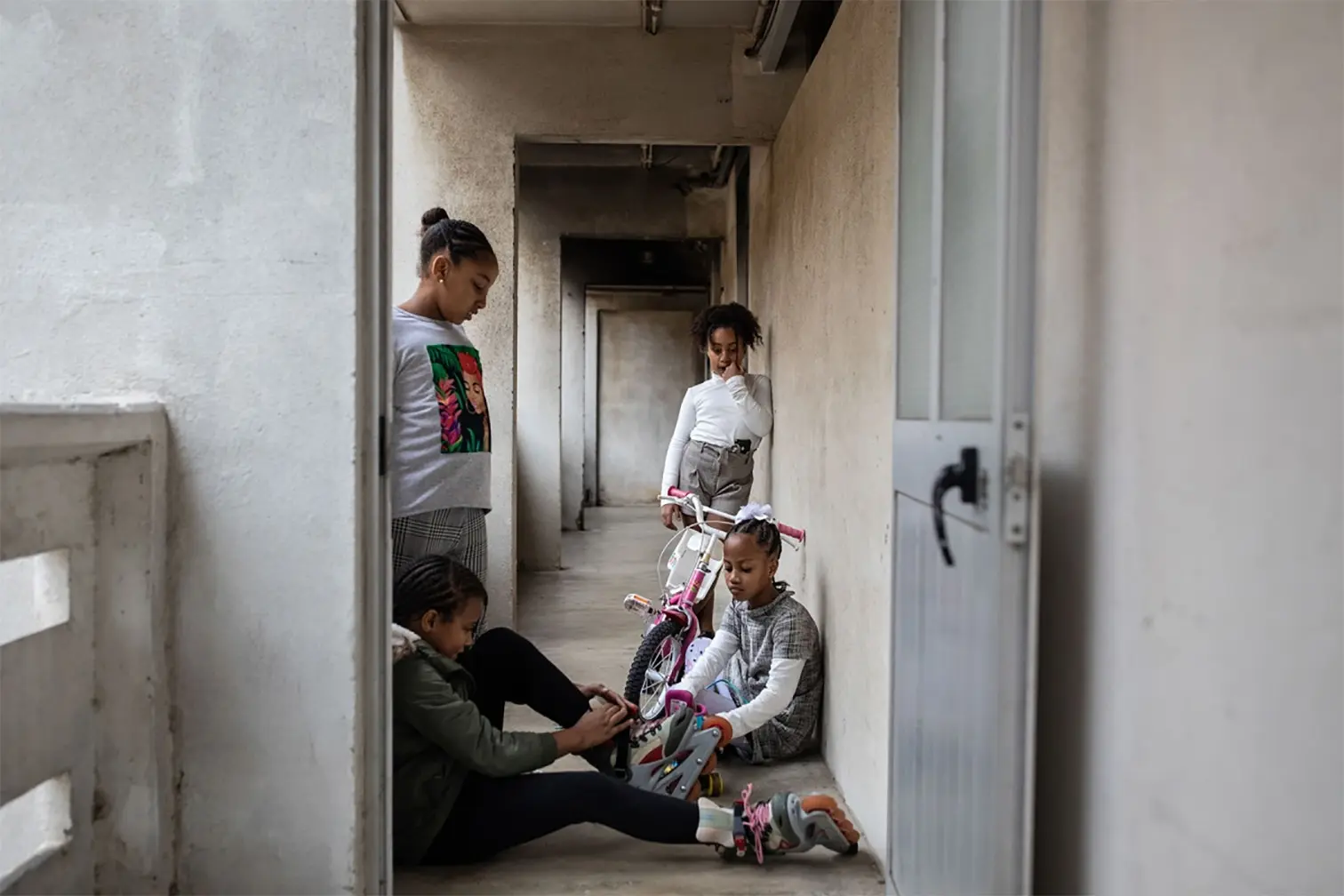 Children in a residential hallway, one sitting on the floor putting on roller skates, others standing nearby with a bicycle.