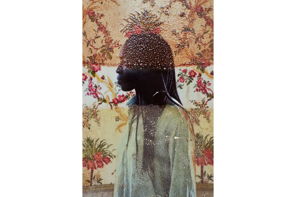 Portrait of a coloured woman in front of a wall with flowers photographed through a water-stained window