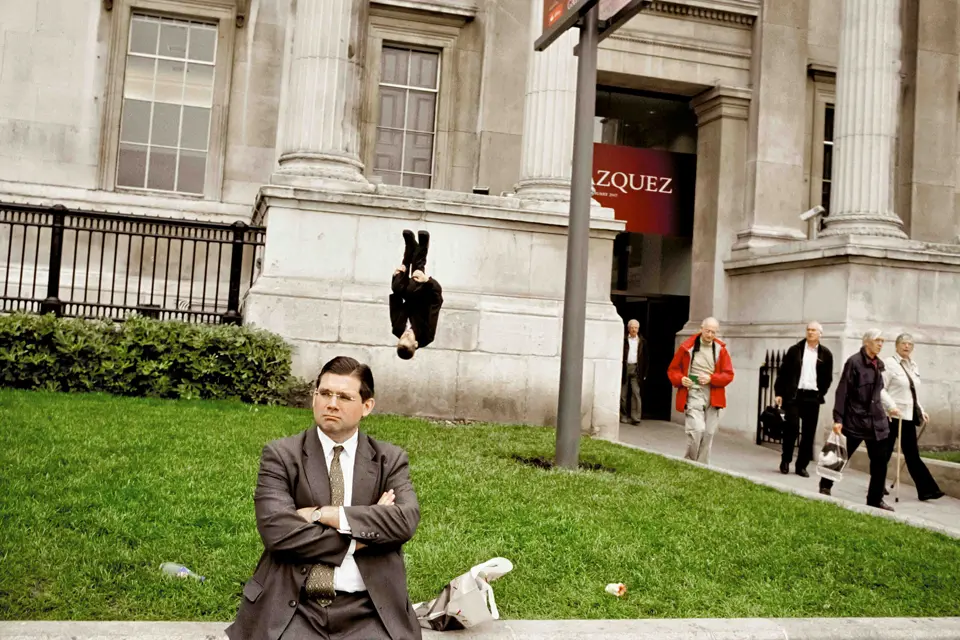 Man in a suit sits on a wall behind him a man in a suit does a somersault