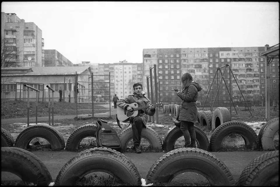 Black and white photograph of a couple on seats made of tires and holding a guitar.