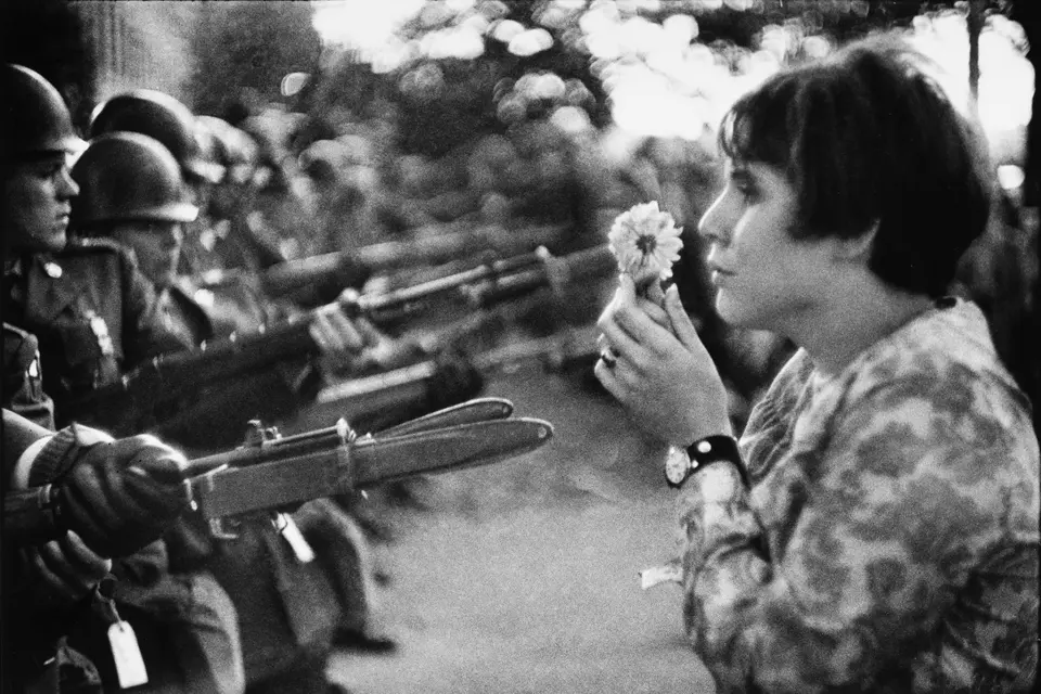 A woman is holding a flower in front of her face and standing in front of armed soldiers.