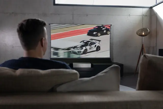 A man sitting on the couch watching a Leica Cine 1 projection showing a car race.