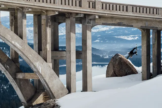 Snowboarder does a one-armed handstand under a bridge on the mountains