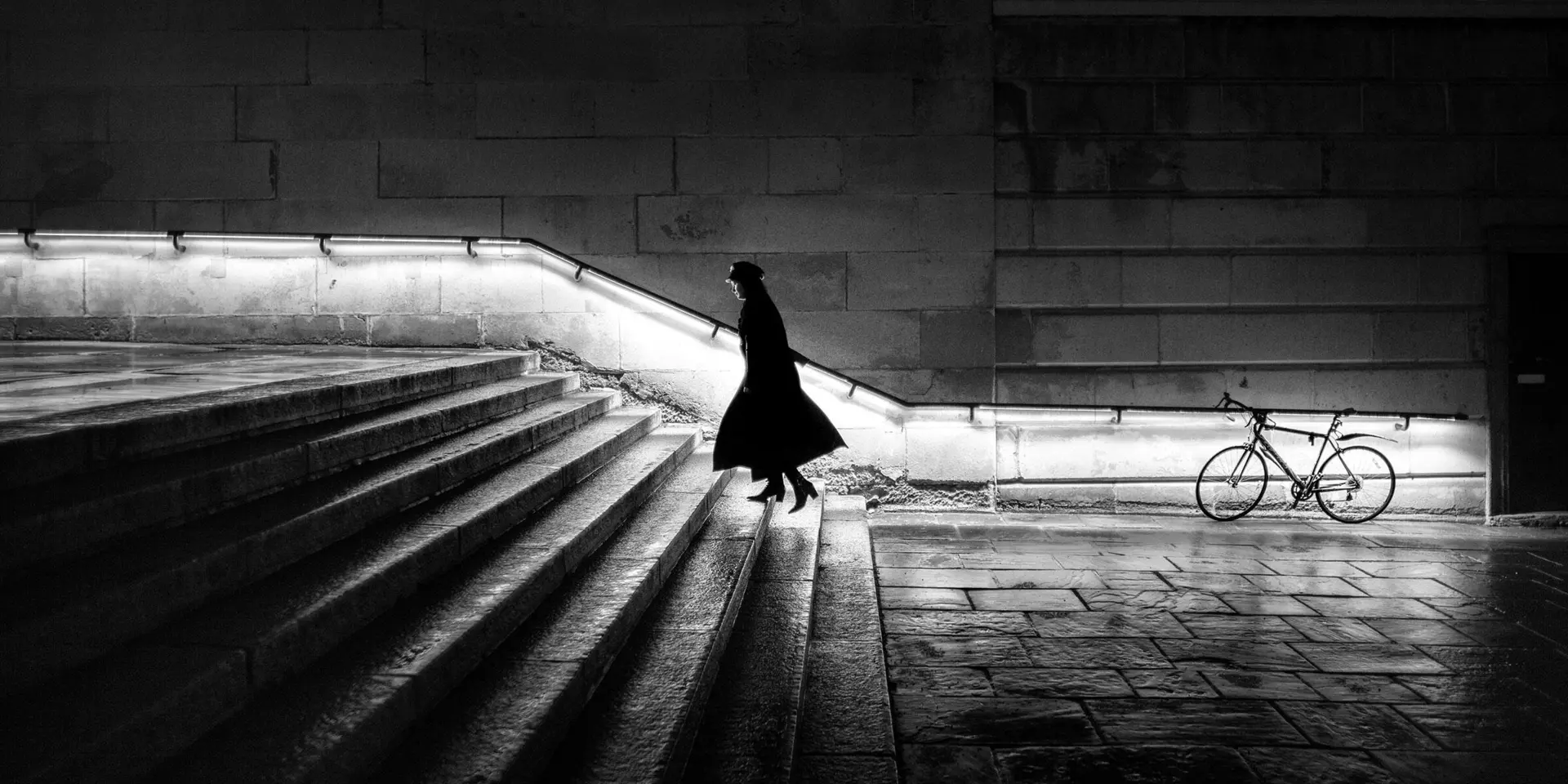 A woman in a long coat is walking up a staircase at night. The banister is illuminated.