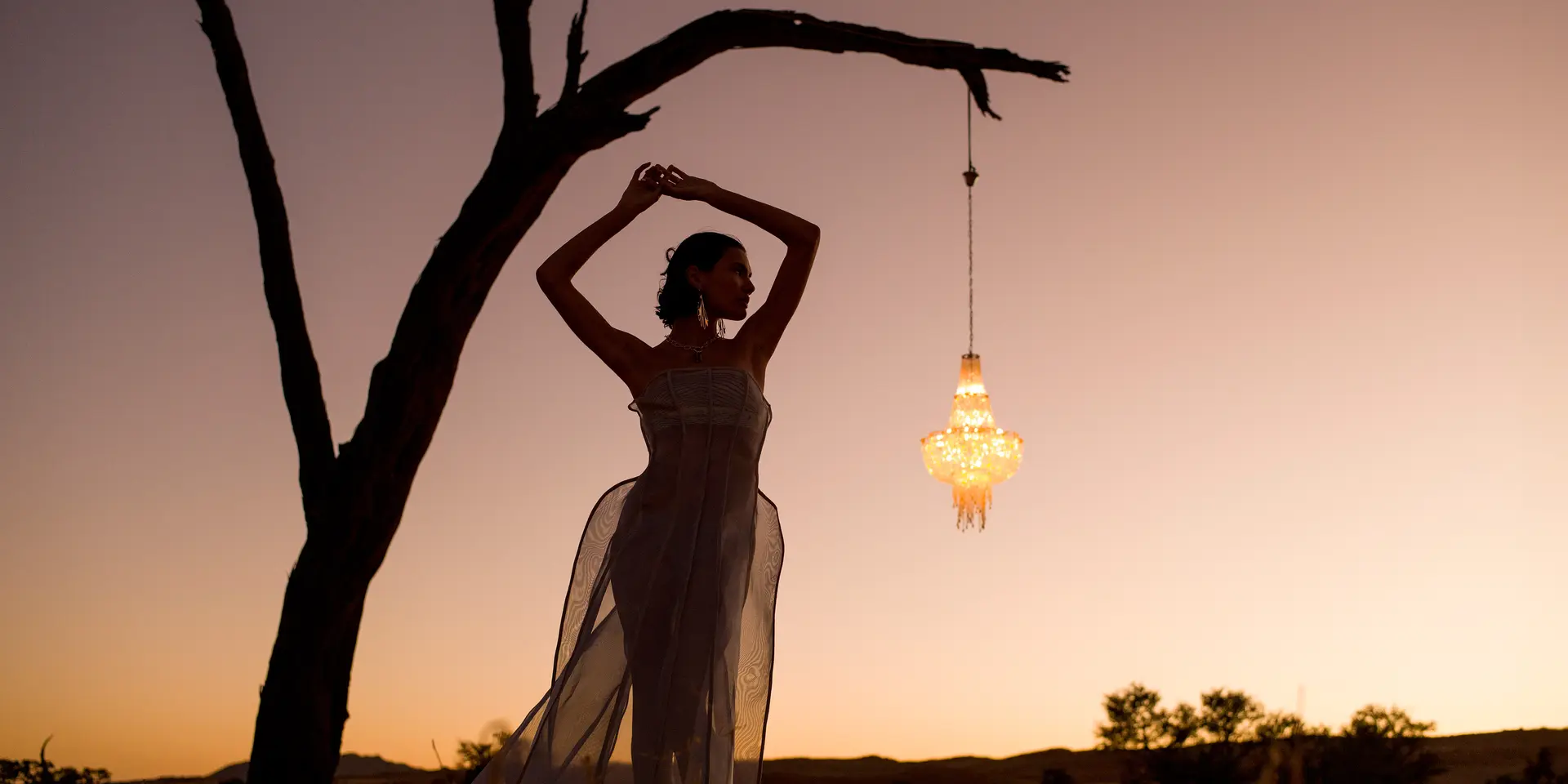 Woman posing under a tree at sunset next to a hanging lantern.