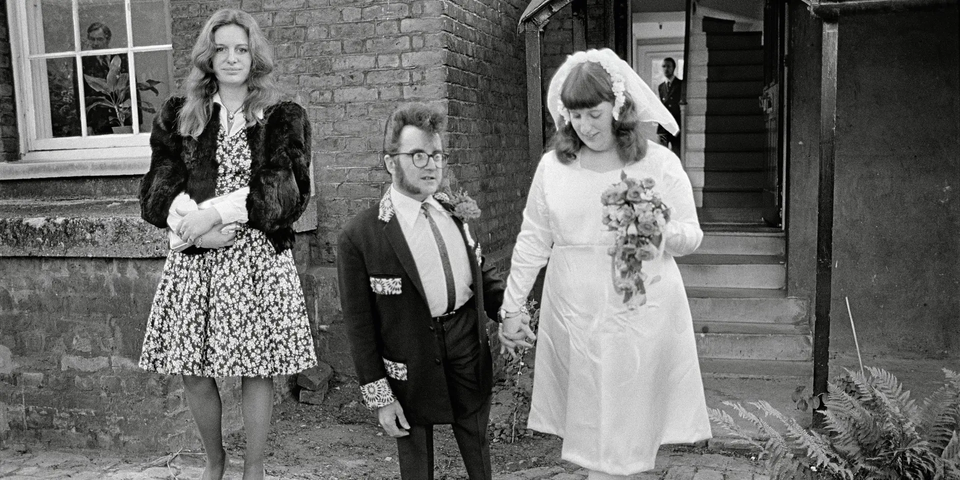 A wedding couple and a woman in a dress stand in front of a building.