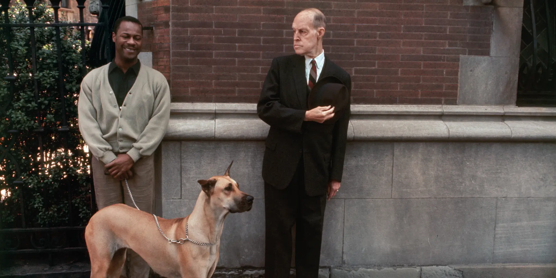 Two men, one with a dog on a lead, stand in front of a brick wall.