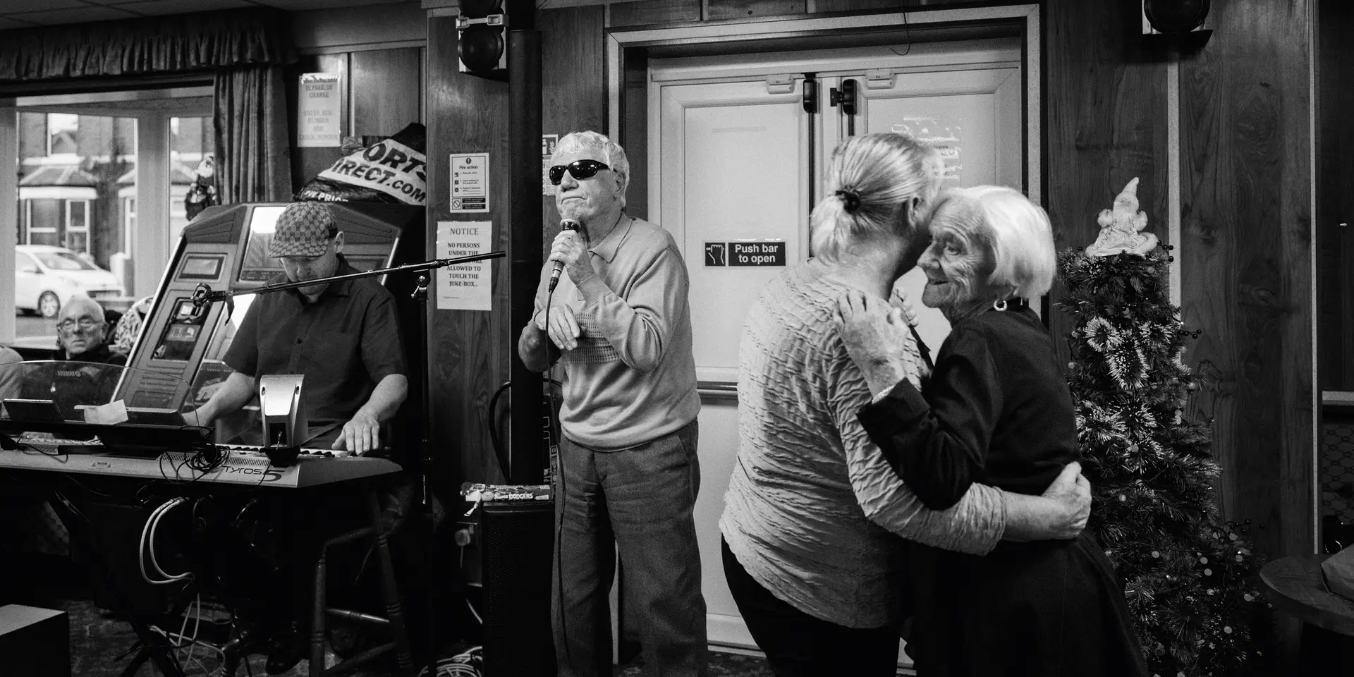 A man sings into a microphone while two older women dance and a man plays the keyboard in the background.