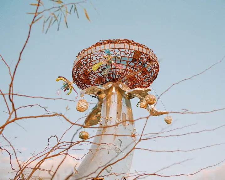 Metallic sculpture with colourful details in front of a blue sky and branches.