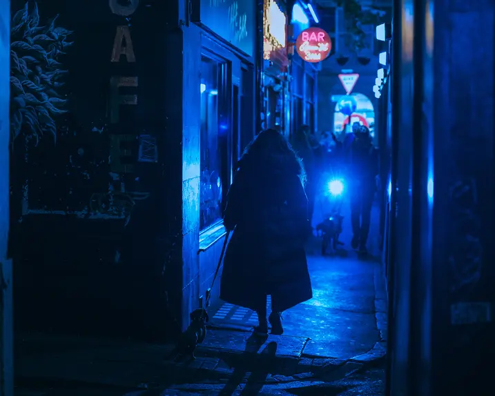 Person walking a dog through a narrow alley at night, illuminated by blue neon lights.