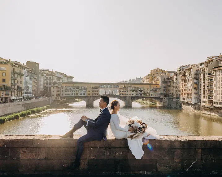A wedding couple sits on a wall overlooking the river, surrounded by old buildings.