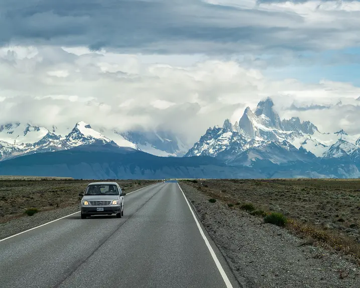 A car is driving on a road with snow-covered mountains in the background.
