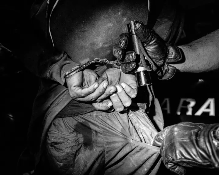 Close-up of a handcuffed person and another person searching his pants pocket with a flashlight.