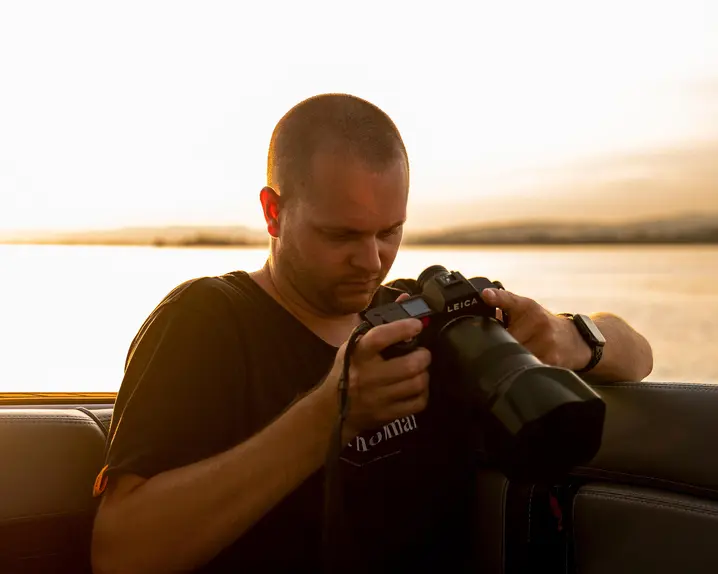 Tino Scherer looking at his camera. In the background is a sea at sunset.