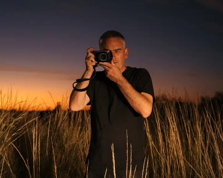 Person in a field with tall grass taking photos with a camera at sunset.