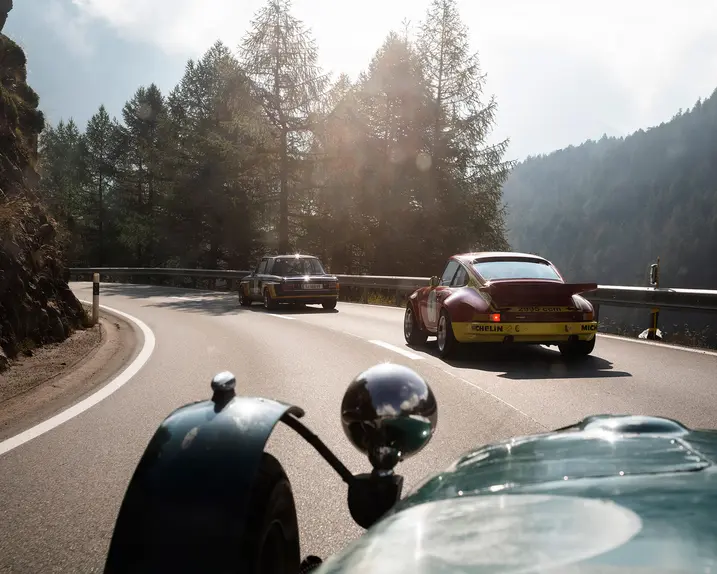 Vintage cars drive along a winding mountain road surrounded by trees and rocks.