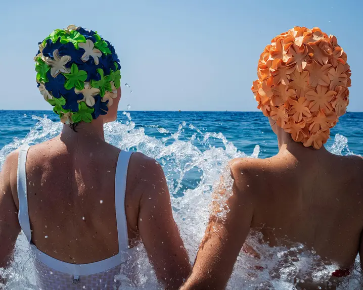 Two people in swimsuits and colorful bathing caps sitting in splashing water in the sea.