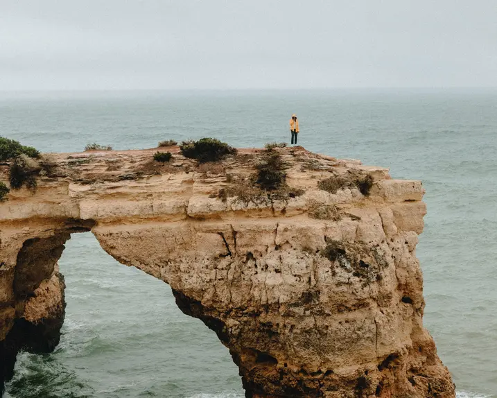 A person is standing alone on a rocky cliff jutting out into the ocean, and it is raining.