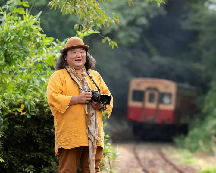 Man with camera stands beside railroad tracks and a train in background.
