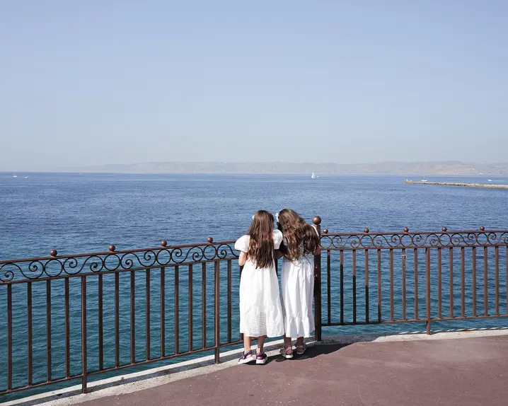 Two girls in white dresses stand at a railing and look to the sea.