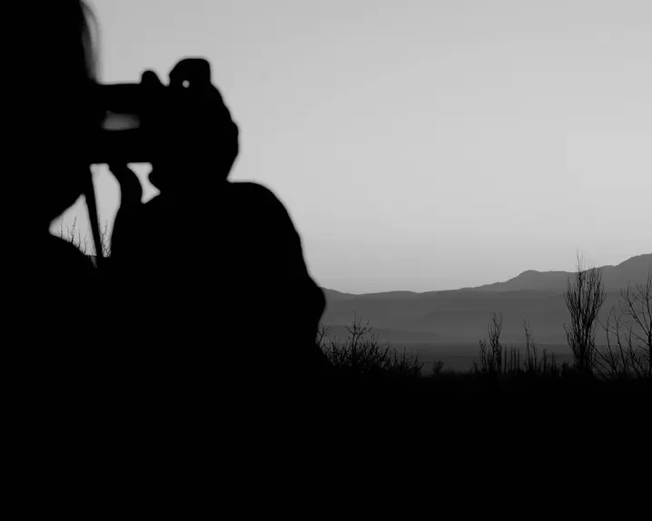 Black-and-white photograph of a person taking a picture of a lake and mountains in the background.