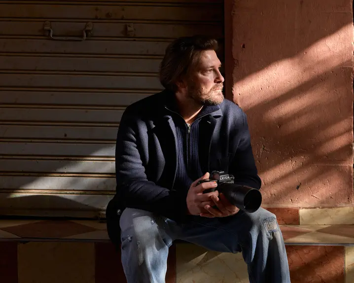 A man sits on a step with a camera in his hands, his face in the sunlight, in front of a closed shutter.