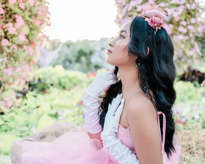 Woman in a pink dress and white gloves is sitting in a rose garden looking to the side.