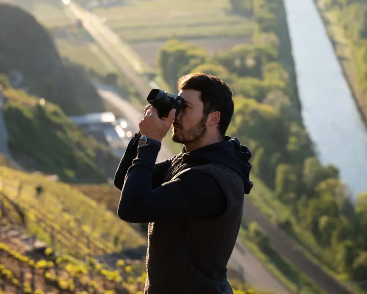 A photographer takes a picture on a sunlit vineyard with a river and trees in the background.