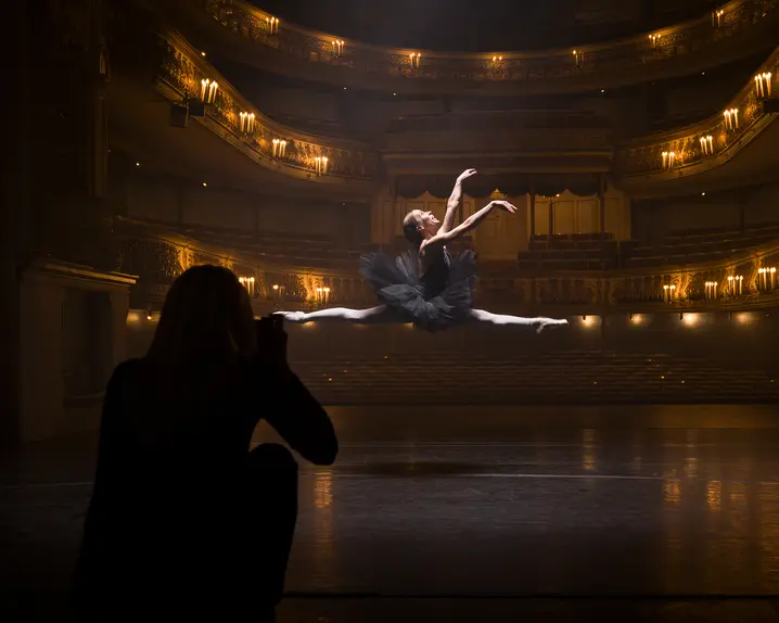 A person in the shade takes a picture of a ballerina jumping in an illuminated theatre.
