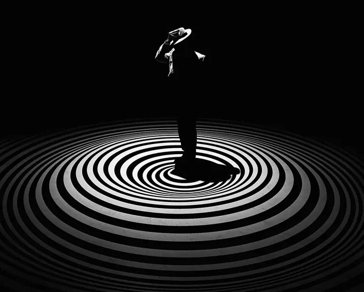 A single person with a hat stands in the centre of concentric circles on a dark background