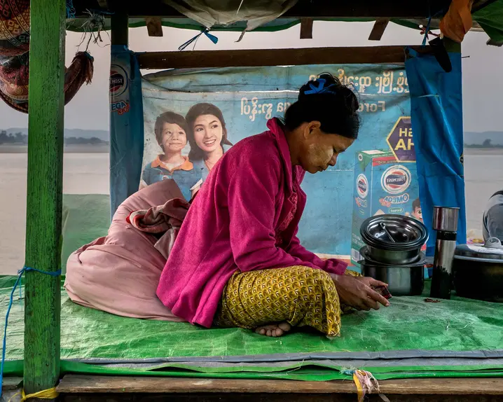 Woman sits on plastic tarp looking at a phone, in background are other people and a river.