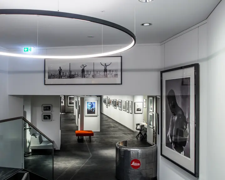 View down a staircase into a long corridor with framed pictures on the wall. At the beginning is a semi-circular metal reception desk with Leica written on it.