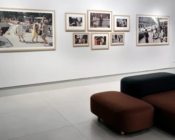 Framed pictures of people in the city hang on a white wall, with black and red upholstered stools in front of them.