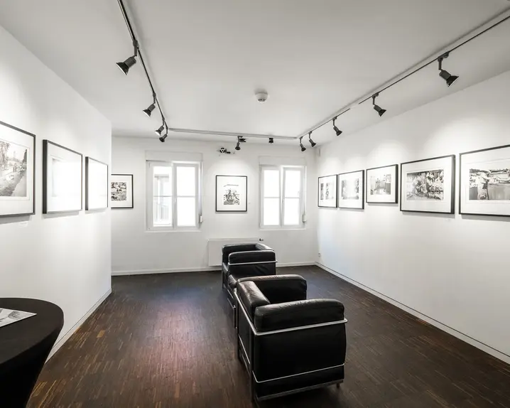 Exhibition room with black and white photographs on the wall and black leather armchairs in the centre of the room.